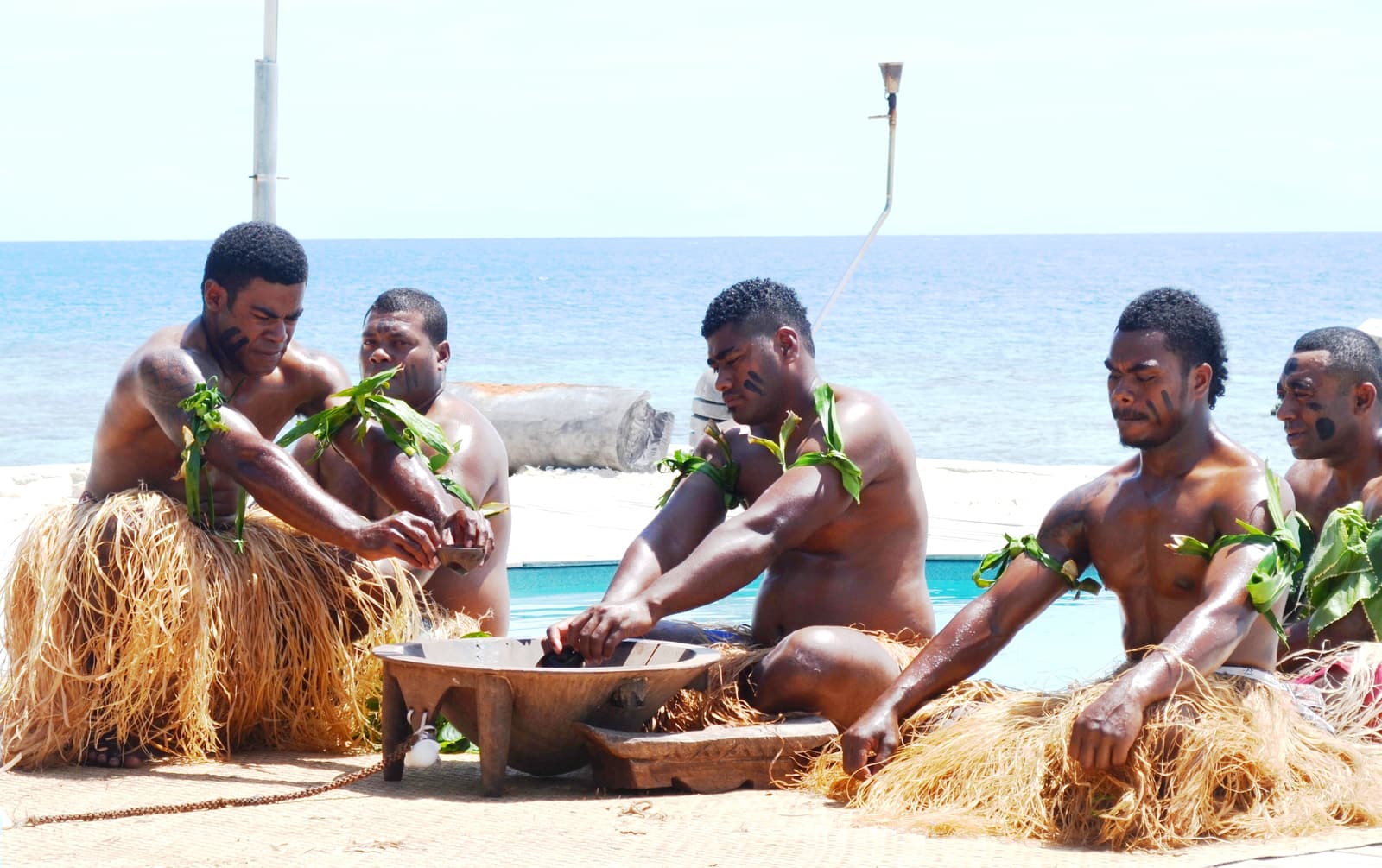 Fijian men in traditional dress preparing kava in a wooden bowl on the beach